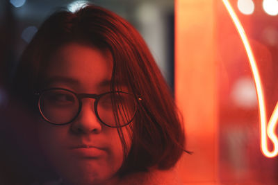 Close-up of thoughtful young woman wearing eyeglasses against illuminated red neon light