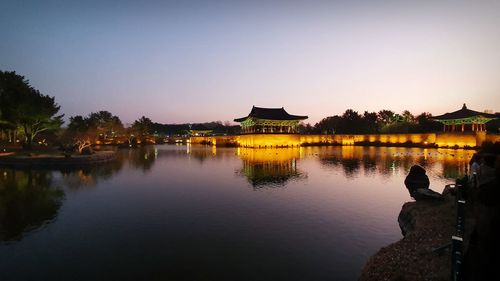 Illuminated building by lake against clear sky during sunset