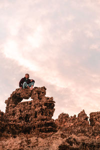 Low angle view of young man sitting on rock against sky