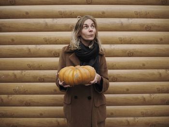 Woman holding pumpkin while standing by wooden wall