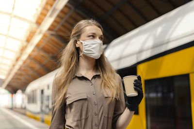 Woman wearing mask holding coffee while standing at railroad station