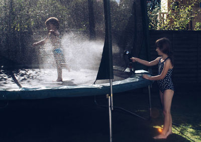 Girl spraying water on her brother standing on trampoline in backyard