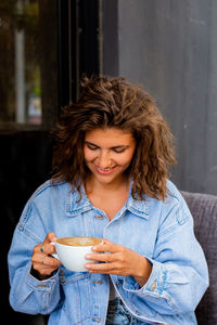 Young charming woman with curly hairstyle holding big mug, having fresh cappuccino and relaxing