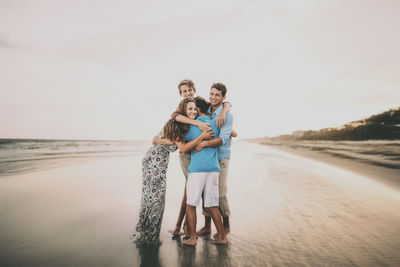 Happy siblings embracing while standing at beach against sky during sunset