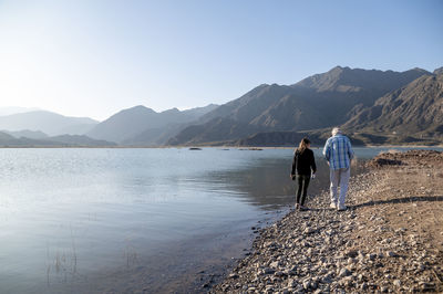 Senior man walking with his granddaughter along the lake shore with mountains.