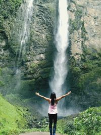 Rear view of woman with arms outstretched standing against waterfall