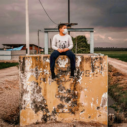 Full length of teenage boy sitting on brick against sky