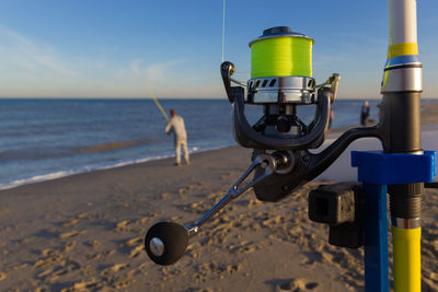 Close-up of fishing rod and reel equipment on beach against sky and sea background