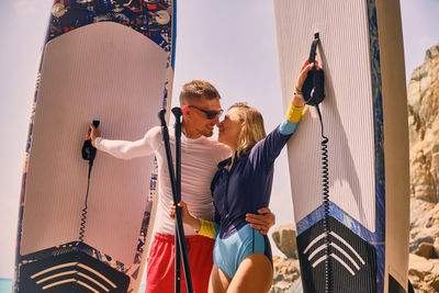 Young active couple stands on a beach kissing and holding stand up paddle boards.