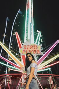 Portrait of young woman standing illuminated amusement park ride at night