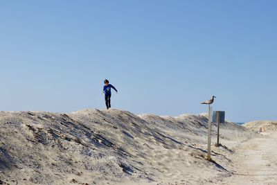 Low angle view of boy standing on dune against clear blue sky