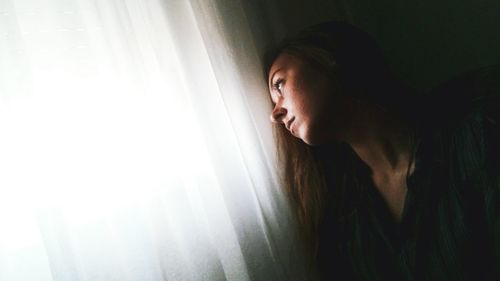 Sad woman looking away while sitting by window curtain at home