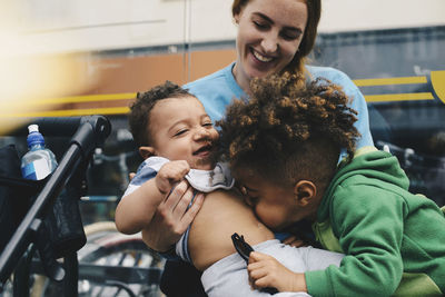 Smiling mother holding baby while brother playing with him at bus stop