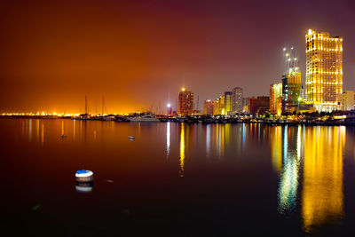 Illuminated buildings by sea against sky at night