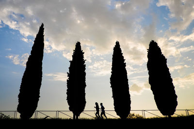 Low angle view of silhouette couple jogging by trees against cloudy sky during sunset