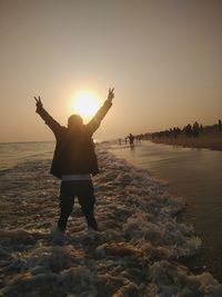 Rear view of man with arms raised standing in sea against clear sky during sunset