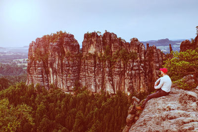 Man hiker with red cap sits on big rock and looks at sharp mountain with tooth peaks