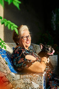 Smiling aged lady in eyeglasses sitting with french bulldog on bench in daylight in yard