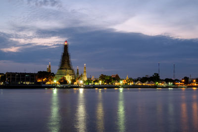 Illuminated buildings at waterfront against cloudy sky