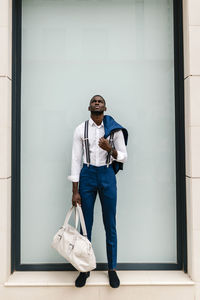 Young male professional holding suit and duffel bag while looking up against window in city