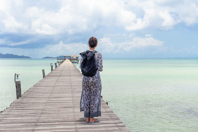 Rear view of woman standing on the wooden bridge looking at sea against sky