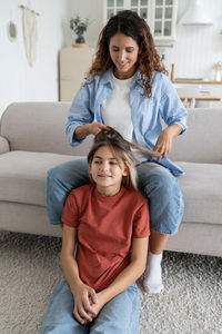 Smiling positive woman sits on sofa combing hair of cheerful school age teen girl dughter and gossip