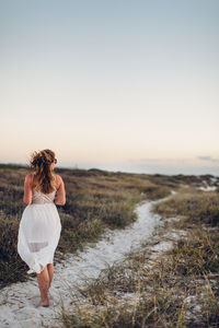 Rear view of young woman walking at beach against clear sky during sunset