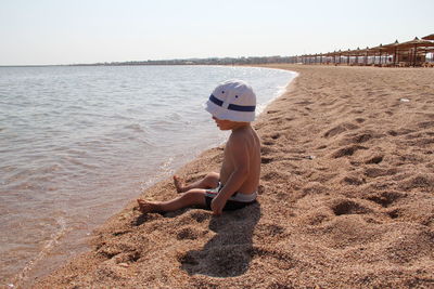 The baby in swimming trunks and panama sits on the beach in the sand by the sea. 