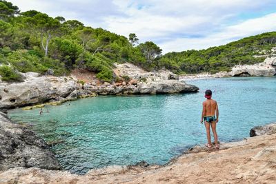 Rear view of shirtless boy looking at bay of water against sky