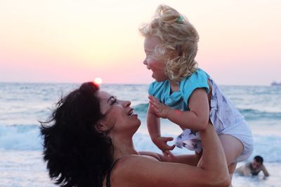 Mother lifting cheerful daughter at beach against sky during sunset