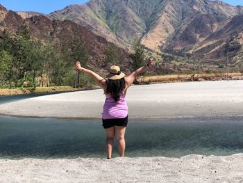 Rear view of woman with arms outstretched standing against mountain at riverbank