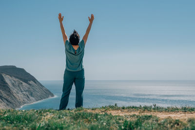 An adult woman stands on a mountain barefoot, raising her hands to the sky and the sun on a sunny