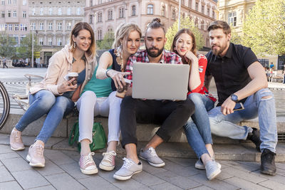 Full length of friends using laptop while sitting at sidewalk in city