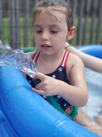 Cute girl holding garden hose while sitting in wading pool at backyard