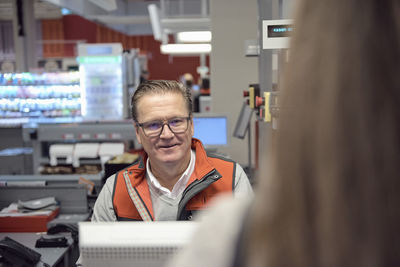 Smiling mature cashier looking at woman at checkout in supermarket