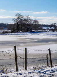 Wooden fence on snow covered field against sky