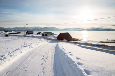 Snow covered road leading towards houses at lakeshore against sky