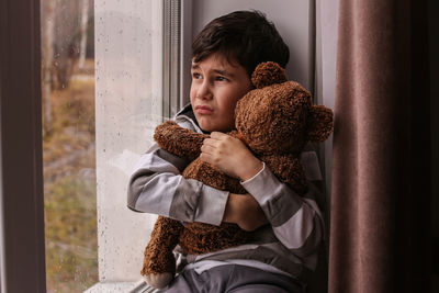 Sad boy looks out the window hugging a teddy bear. bad weather mood, depression