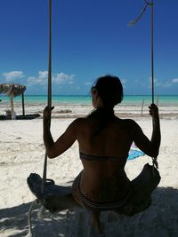 Rear view of woman wearing bikini sitting on swing at beach on sunny day