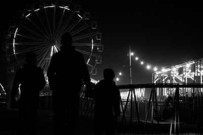 Silhouette people against illuminated ferris wheel against sky at night