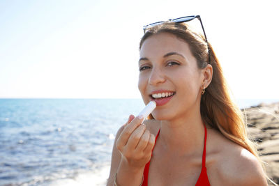 Young woman smiling at camera applying sun protection on her lips on the beach