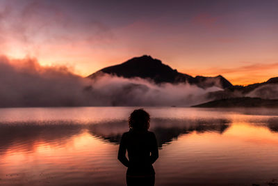 Back view of lonely unrecognizable woman in outerwear looking away thoughtfully while relaxing near calm lake water reflecting mountains and cloudy sky at sunset time