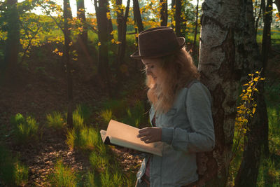 Young woman reading book while standing by trees in forest