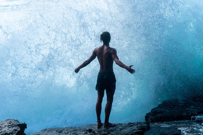 Rear view of shirtless man standing on rocks against waves