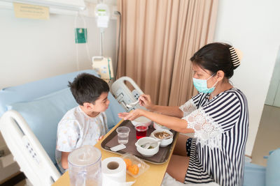 Side view of mother feeding son while sitting on bed in hospital
