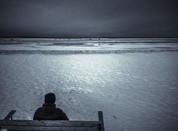 Rear view of boy sitting on bench against snow covered beach