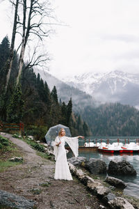 A young woman in a white lace wedding dress stands in the rain among the sea and mountains