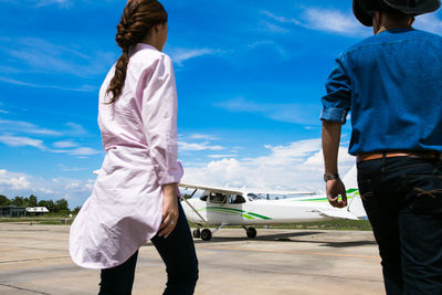 Rear view of couple walking towards airplane on runway against blue sky