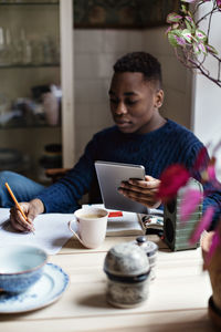 Teenage boy holding digital tablet while writing on book during homework at home