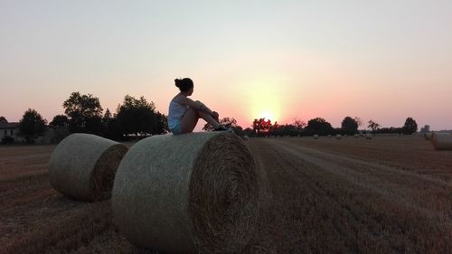 Full length side view of teenage girl sitting on hay bale at farm against sky during sunset